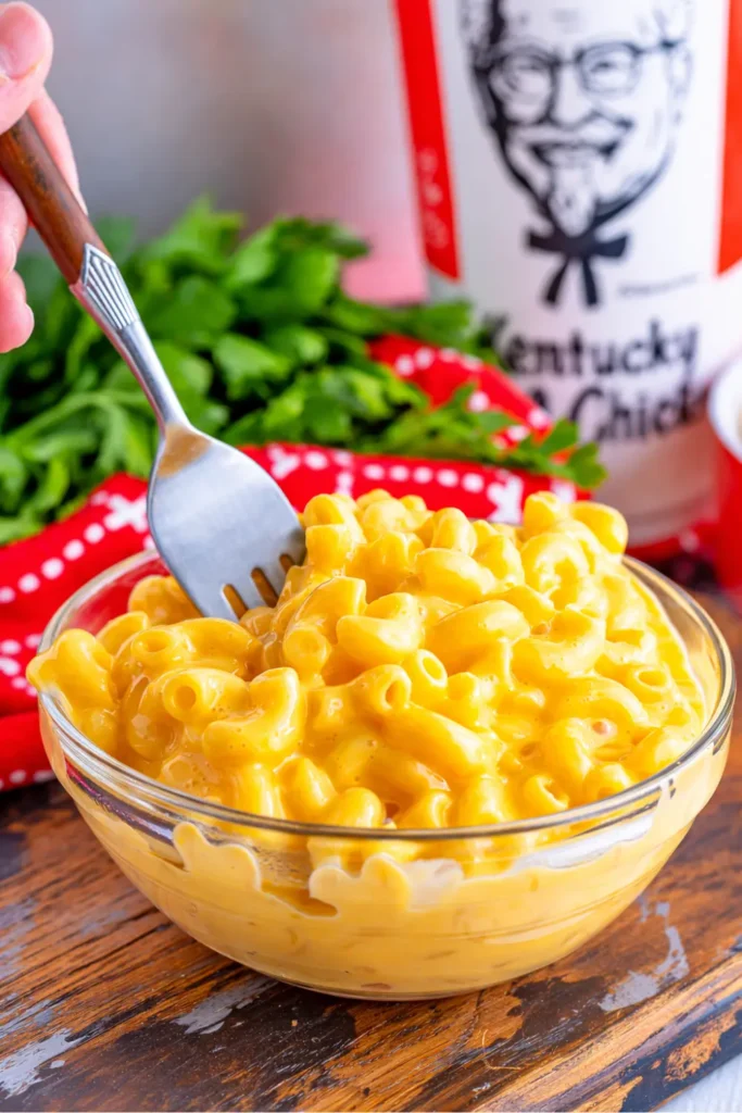 Copycat KFC Mac and Cheese Recipe, Calories & Nutrition Guide 2026 3 A bowl of creamy KFC Mac and Cheese with elbow pasta, rich yellow cheese sauce, and a fork lifting a portion, with a blurred KFC container in the background.