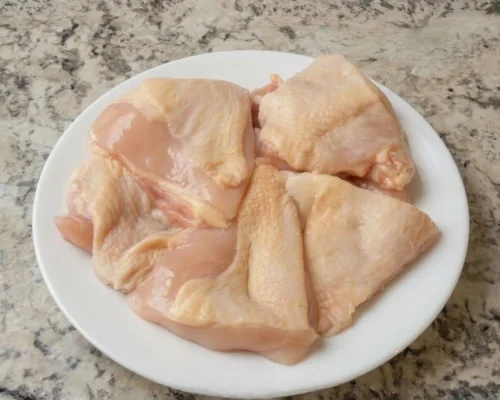 KFC Nashville Hot Tenders Combos Price & Calories 2026 7 Four raw, skin-on chicken thighs on a white plate, the starting point for making KFC Nashville Hot Tenders at home.